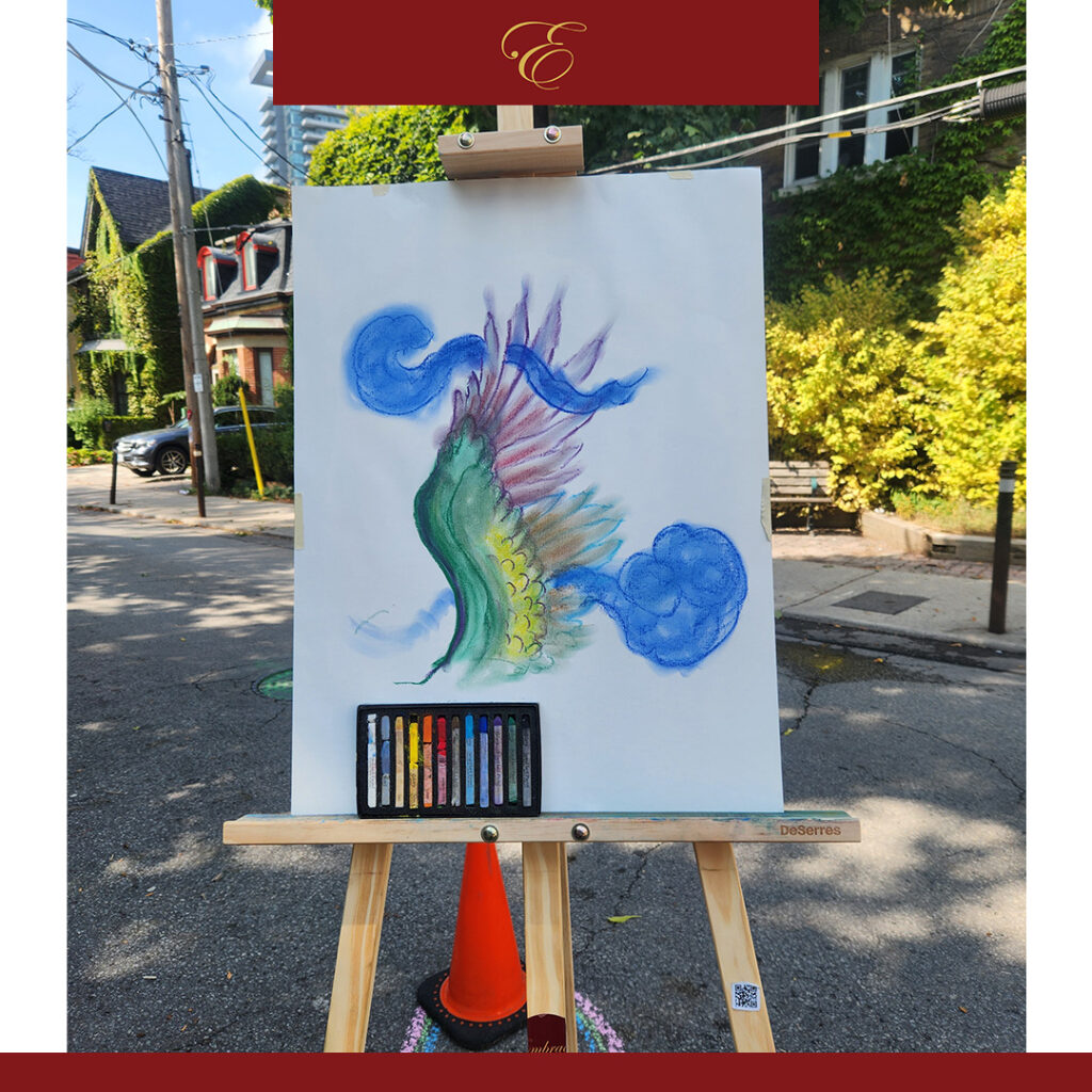 The image features an artwork resting on an easel, placed outdoors on a quiet, sunlit street surrounded by greenery. The artwork itself is an abstract, imaginative piece, featuring a central, flowing form that resembles a mystical creature or wave of energy. Its body is a mix of vibrant greens and yellows, with intricate, scale-like textures transitioning into soft, feathered wings or fins in shades of pink, blue, and purple. Surrounding this figure are two cloud-like, swirling shapes in a rich blue, giving the impression of wind or water in motion. Below the artwork, a small set of colorful pastel crayons rests at the base of the easel, indicating that the piece is still in progress.