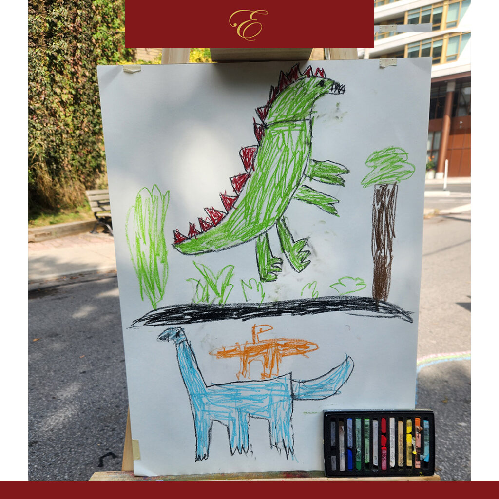 A child’s colorful drawing on display on an easel outdoors, featuring two dinosaurs in a vibrant green and blue jungle setting. The drawing is done with bright crayons, with green and red spiked dinosaurs among plants, trees, and grass, and smaller, differently colored dinosaurs below. The crayons used for the artwork are placed at the bottom of the page.